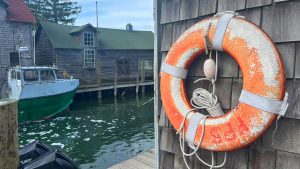 The right side of the image features a worn life ring hanging on a wall made of wooden shakes. The left side is a narrow body of water with a fishing boat in it, and a fishing shack on the far side.