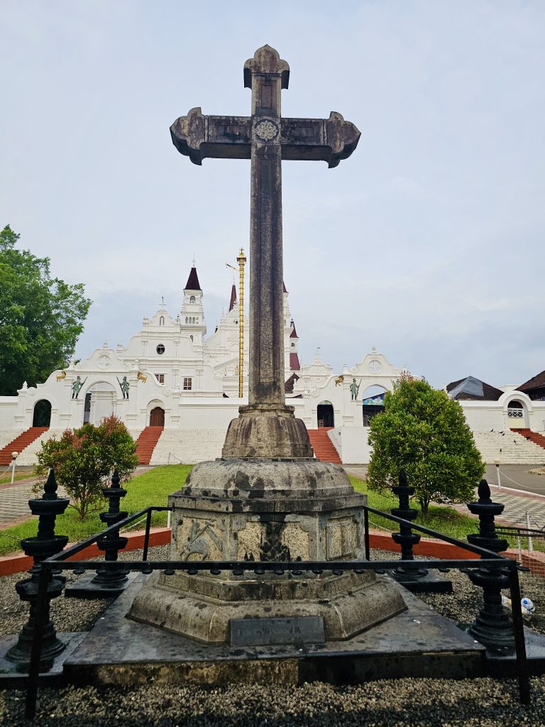 A large stone cross stands prominently in front of a beautiful white church. The landscaped courtyard, steps, and church architecture create a strong and peaceful composition. Location: Ramapuram, Kottayam, Kerala.