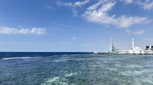 Wide view of the deep blue and turquoise Red Sea off Jeddah, with shallow reef visible under the water and the white floating mosque and the glass sail-shaped tower of Jeddah Yacht Club on the distant shoreline to the right.
