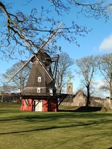 Historic red wooden windmill with large sails standing on a grassy lawn at Kastellet in Copenhagen, framed by bare tree branches under a blue sky.
