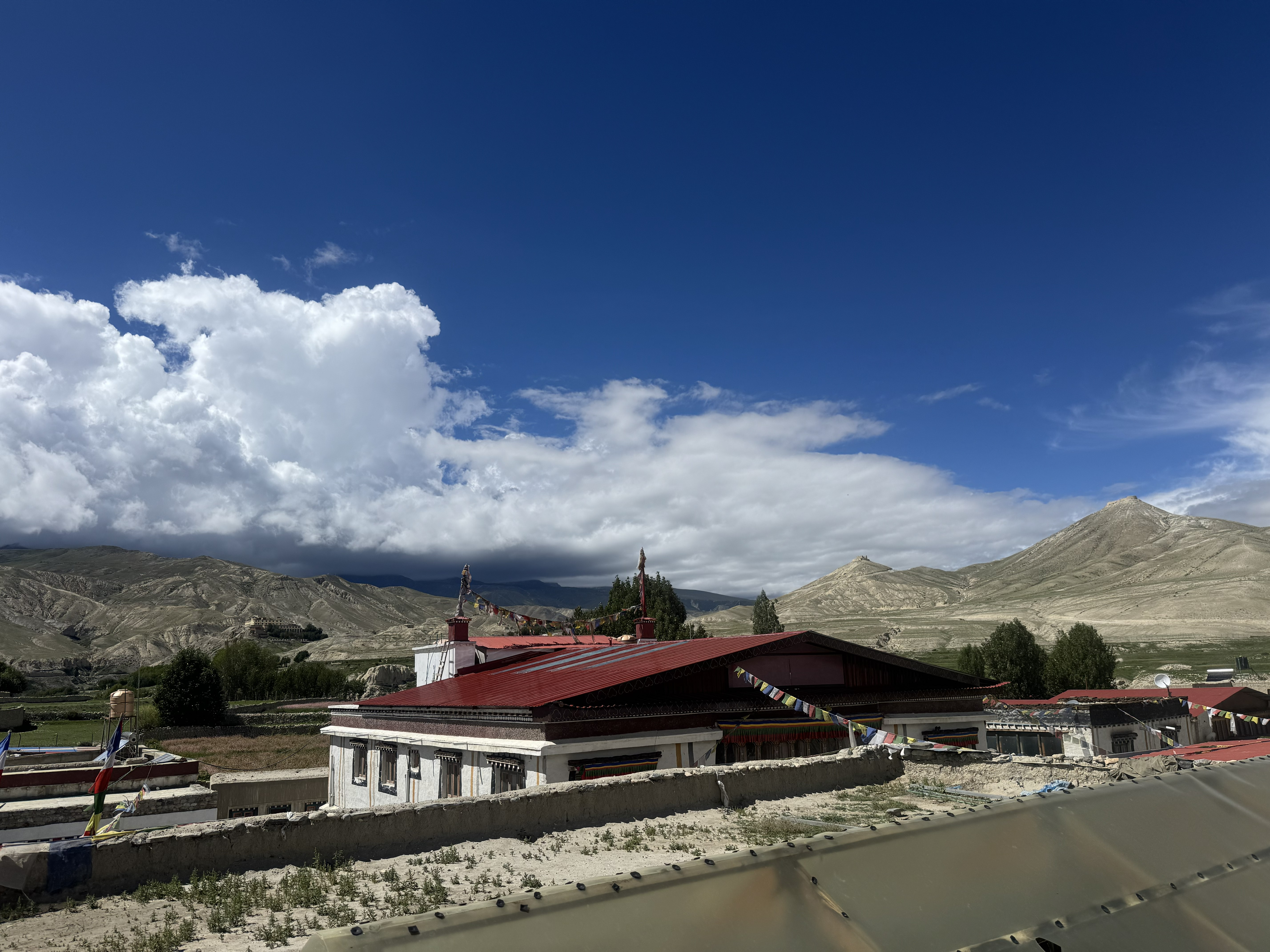 A traditional red-roofed building adorned with prayer flags, set against a backdrop of barren, light-brown mountains beneath a bright blue, cloud-dotted sky.