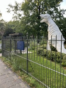 White dinosaur statue in an urban park surrounded by greenery behind a fence.
