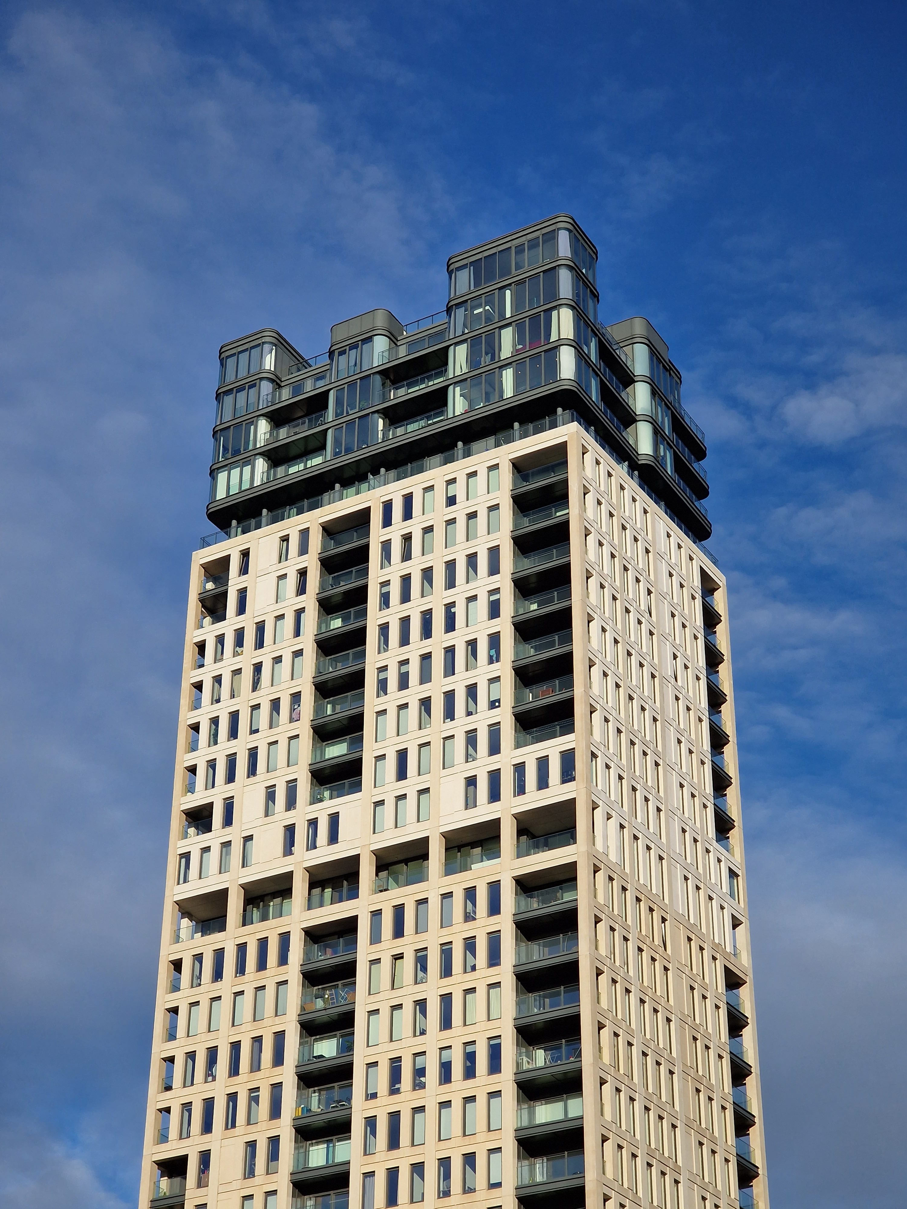 Vertical low-angle shot showing the upper floors and glass penthouse of a modern beige residential tower against a blue sky.