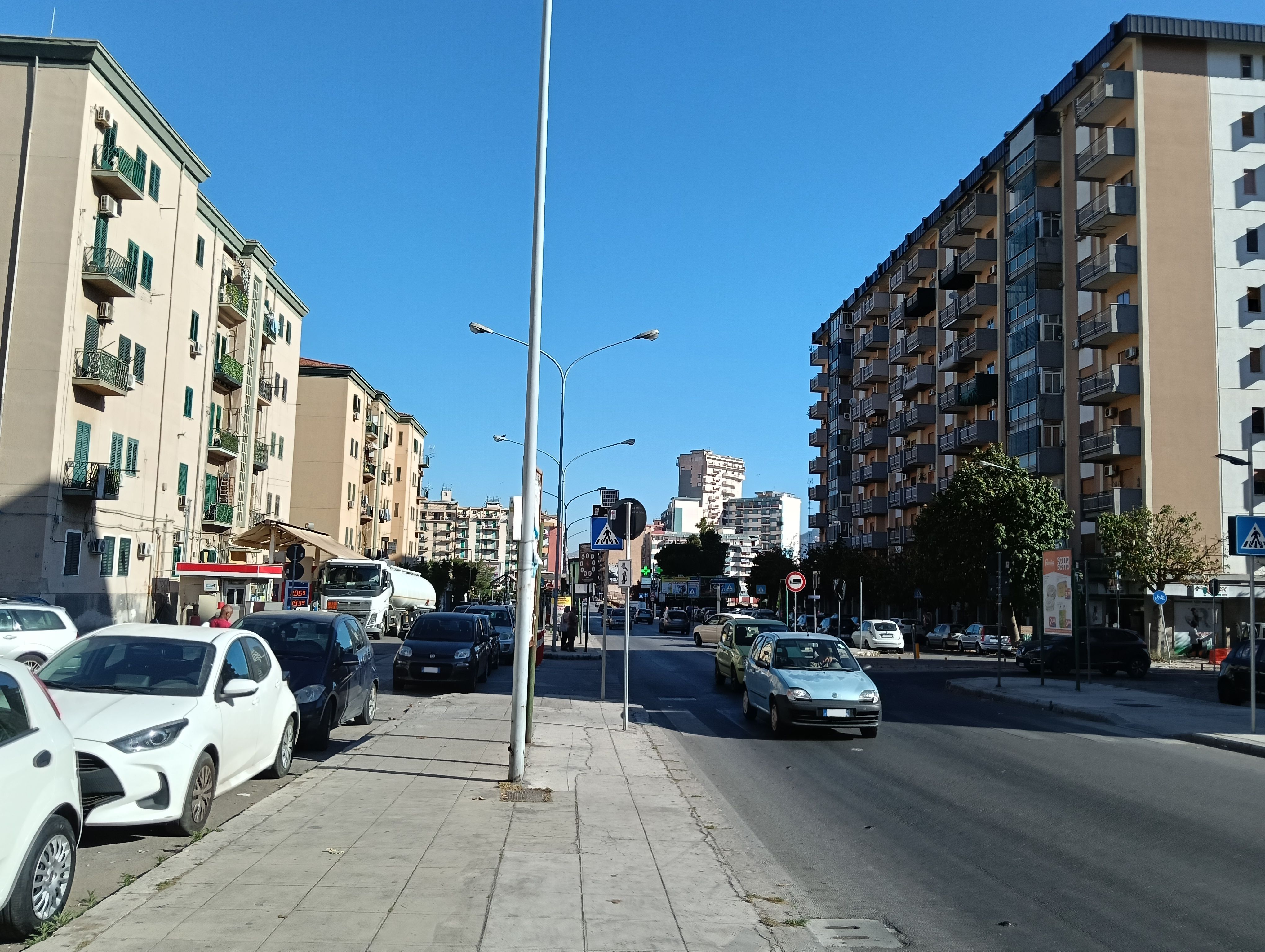 Concrete pathway with lamps along a road with a couple of cars and buildings with a clear sky at the bottom.