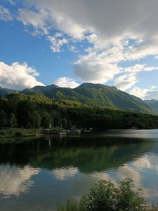 Scenic alpine lake reflecting green forested mountains and blue sky with fluffy white clouds.