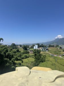 A wide landscape showing green agricultural terraced fields, rural houses, and surrounding hills stretching towards mountains under a clear sky.