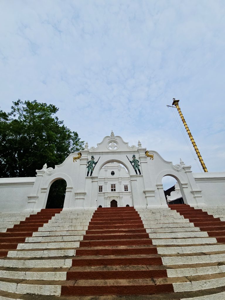 A wide-angle view of the church entrance with red-and-white steps, decorative arches, statues, and a tall golden flagpole. The upward perspective gives the historic-looking façade a dramatic appearance. Location: Ramapuram, Kottayam, Kerala.