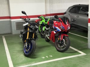 Three motorcycles parked closely together in an indoor garage with a green floor, featuring a black naked bike, a bright green sportbike, and a red sportbike.
