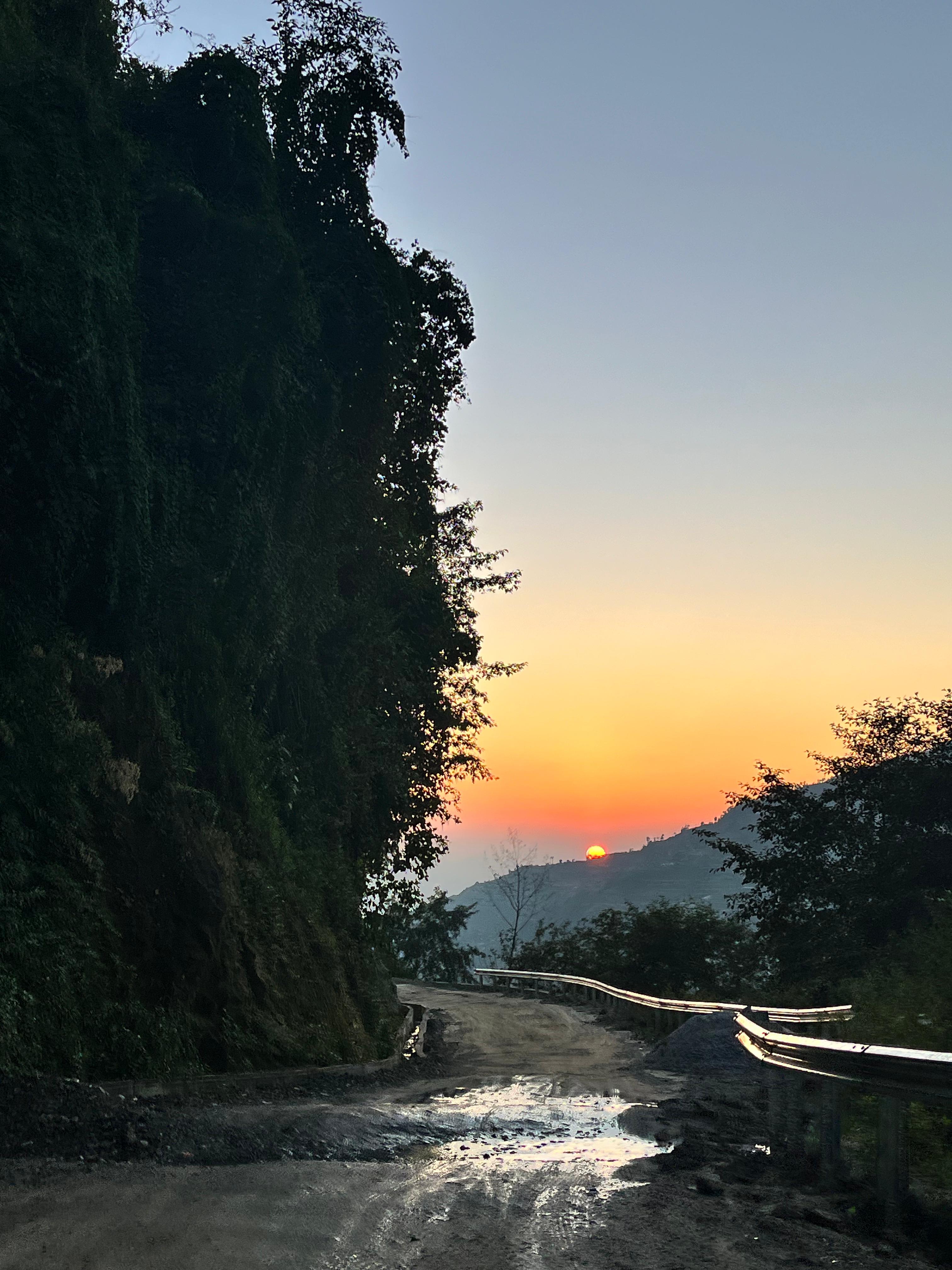 Winding wet dirt road through dense greenery at sunset, with a hillside and warm orange sky in the background.