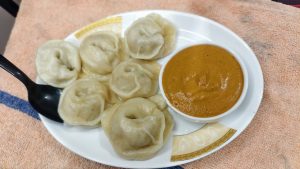 A white ceramic plate served with seven freshly steamed meat momo dumplings arranged next to a small bowl of thick orange dipping sauce and a black plastic spoon.