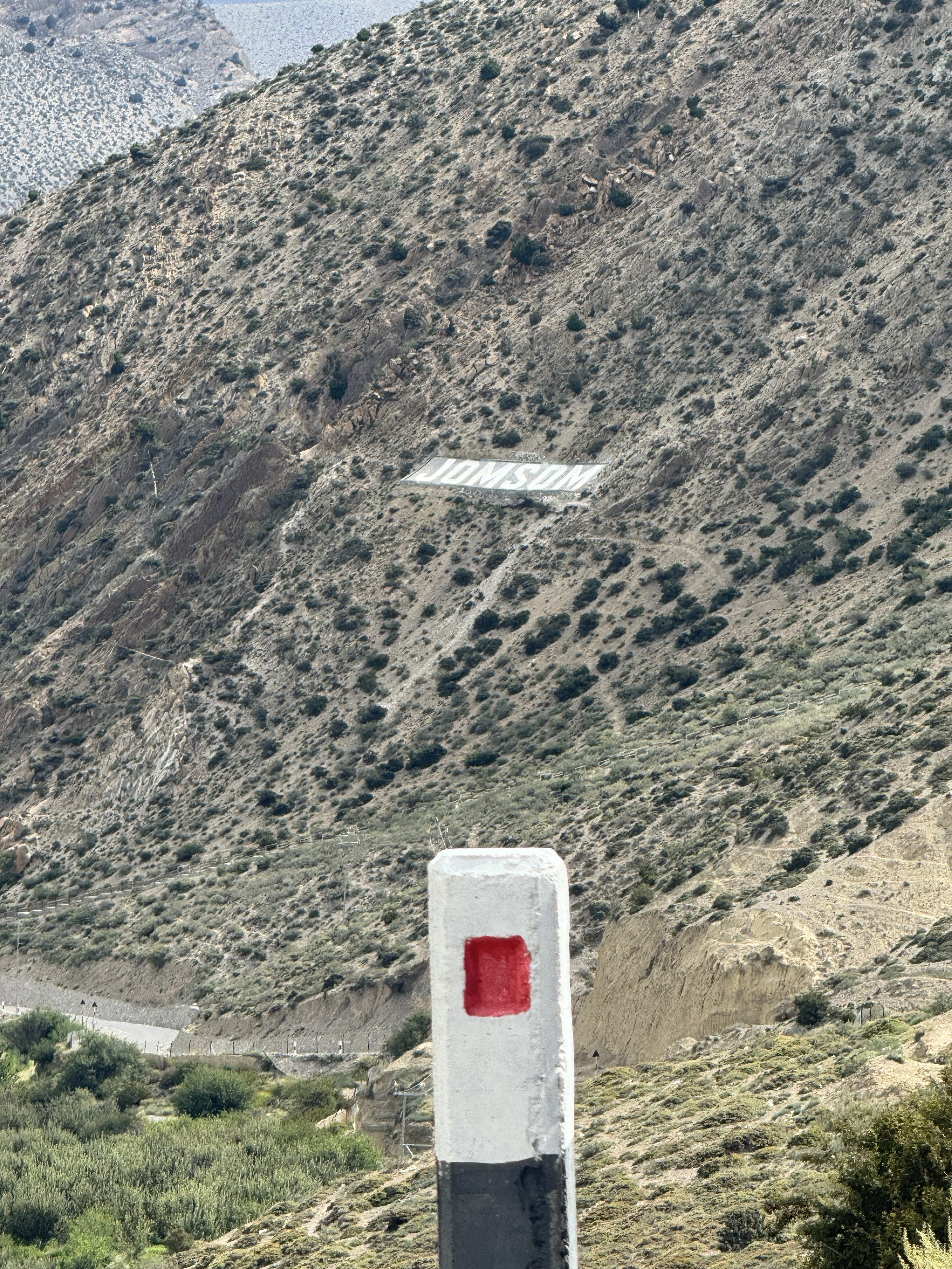 A white milestone post with a red square in the foreground, set against a steep, arid mountain slope featuring a large white "JOMSOM" landmark sign.