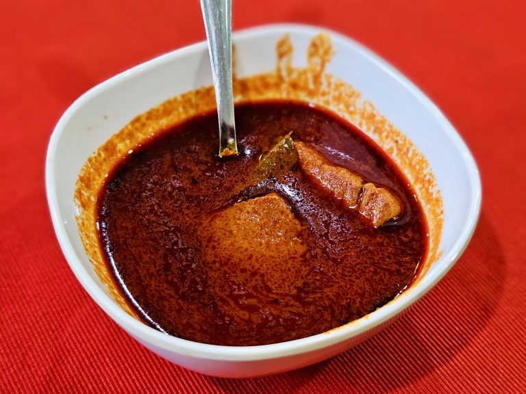 A bowl of rich red-brown fish curry sits on a red tablecloth, with visible spices and ingredients adding texture. The warm colors give the photograph a distinctly appetizing appearance. Location: Ayamkudy, Kottayam, Kerala.