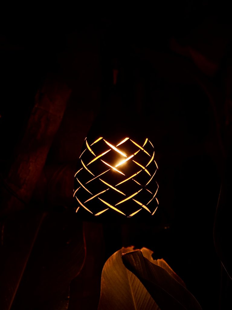 A glowing decorative lamp with an intricate woven pattern creates a warm, artistic scene against a dark background. The strong contrast beautifully highlights the light and surrounding shapes. Location: Edavannappara, Malappuram, Kerala