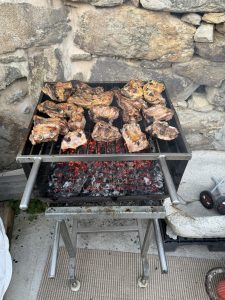 Outdoor metal barbecue on wheels grilling various cuts of meat and ribs over hot coals, with a rustic stone wall in the background.