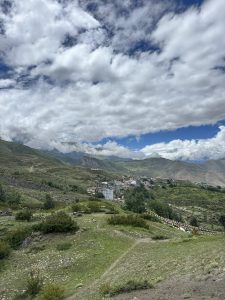 Mountain valley with a remote village, colorful prayer flags, and green slopes under a cloudy blue sky.