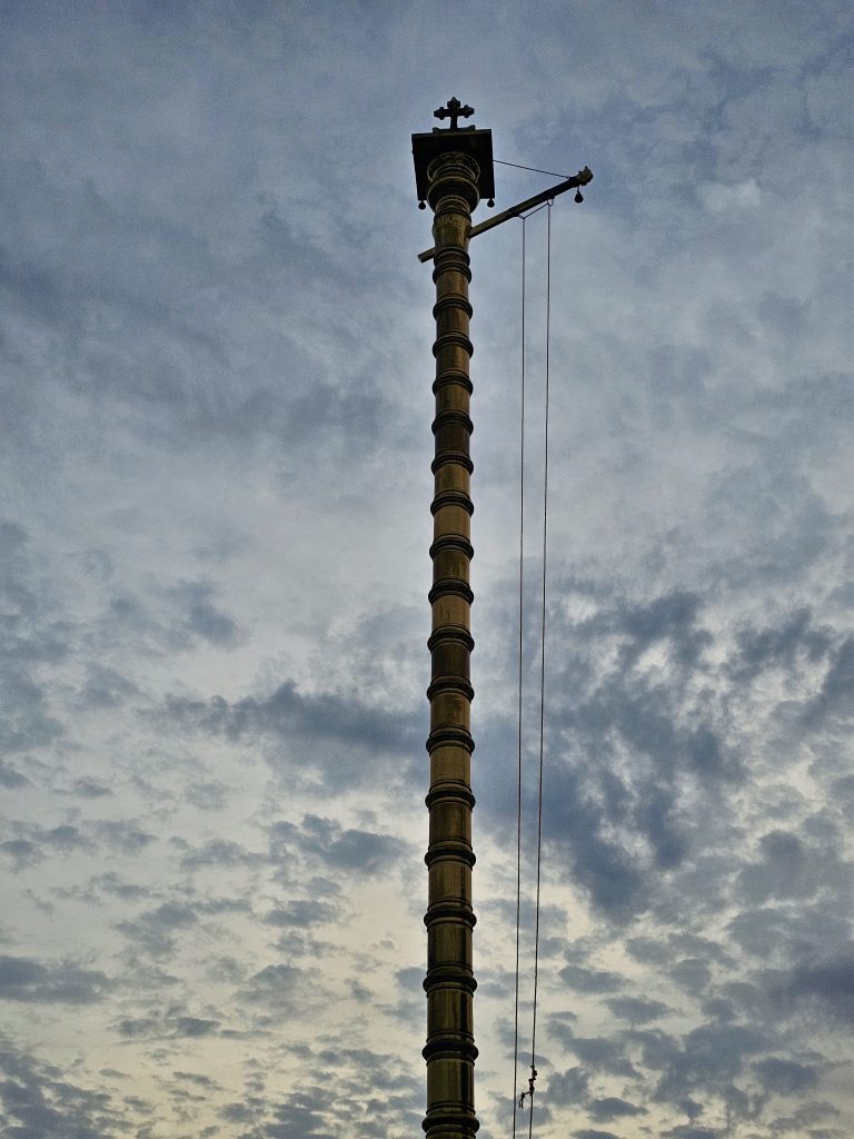 A tall golden flagpole rises dramatically against a cloudy evening sky. The upward composition highlights its detailed structure and creates a strong architectural photograph. Location: Ramapuram, Kottayam, Kerala.