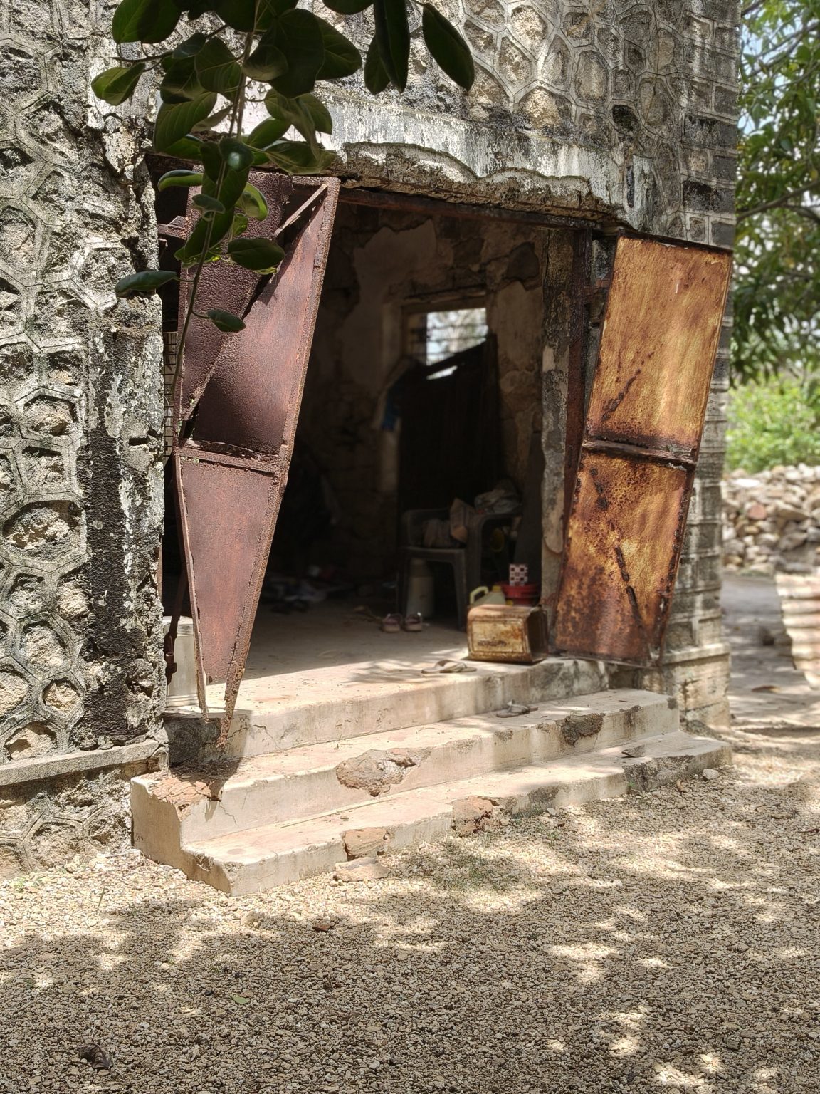 Old weathered stone building with rusted metal doors, uneven steps, and leafy branches overhead.