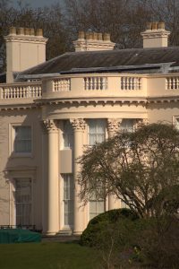 Telephoto shot of a cream-colored classical building exterior featuring Corinthian columns, a curved portico, decorative balustrade, and chimney stacks.