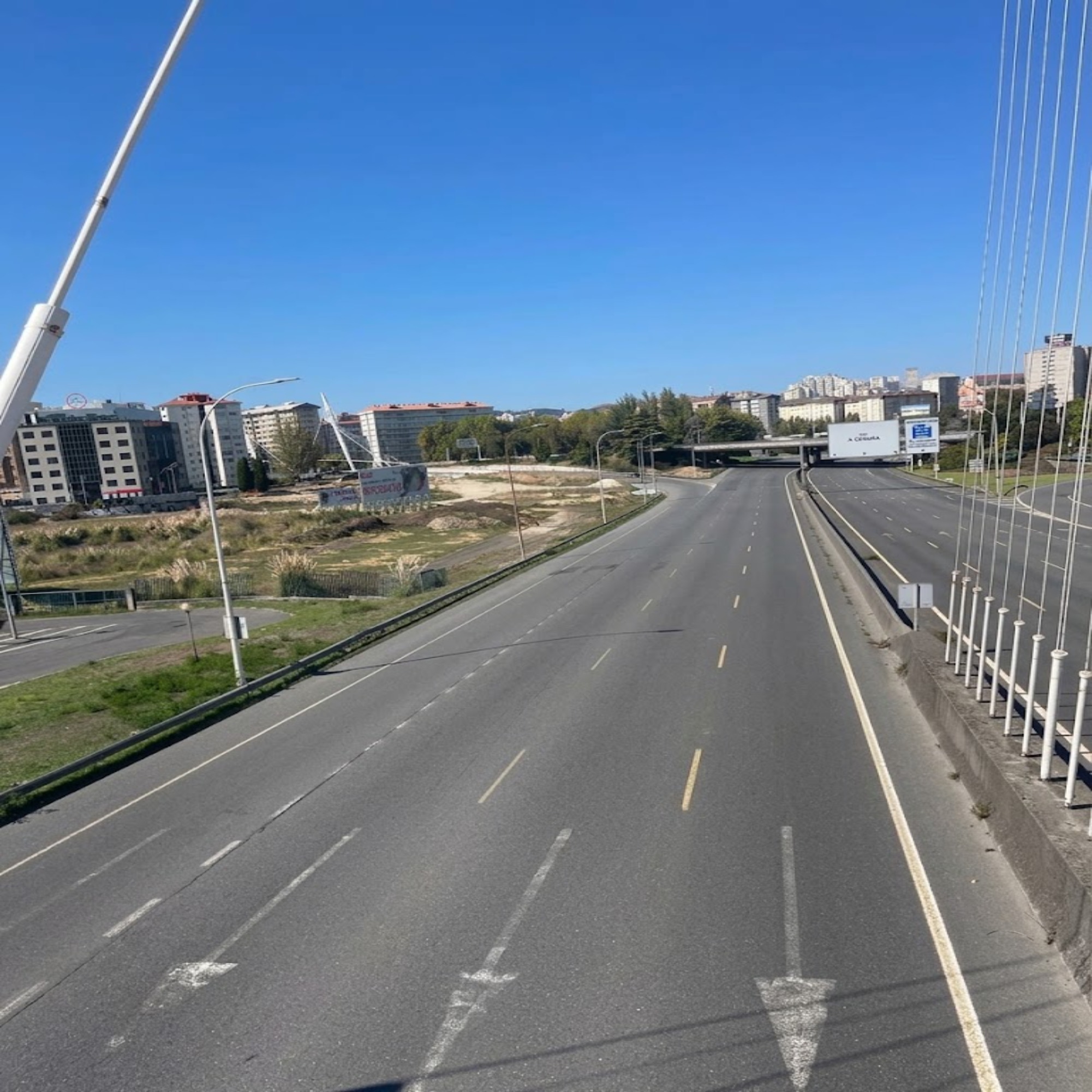 Wide, empty road with grassy areas, tall buildings, and a large billboard under a clear blue sky.
