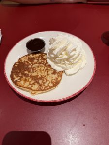 Golden pancake with whipped cream and a small container of dark chocolate syrup on a white plate with a red rim.