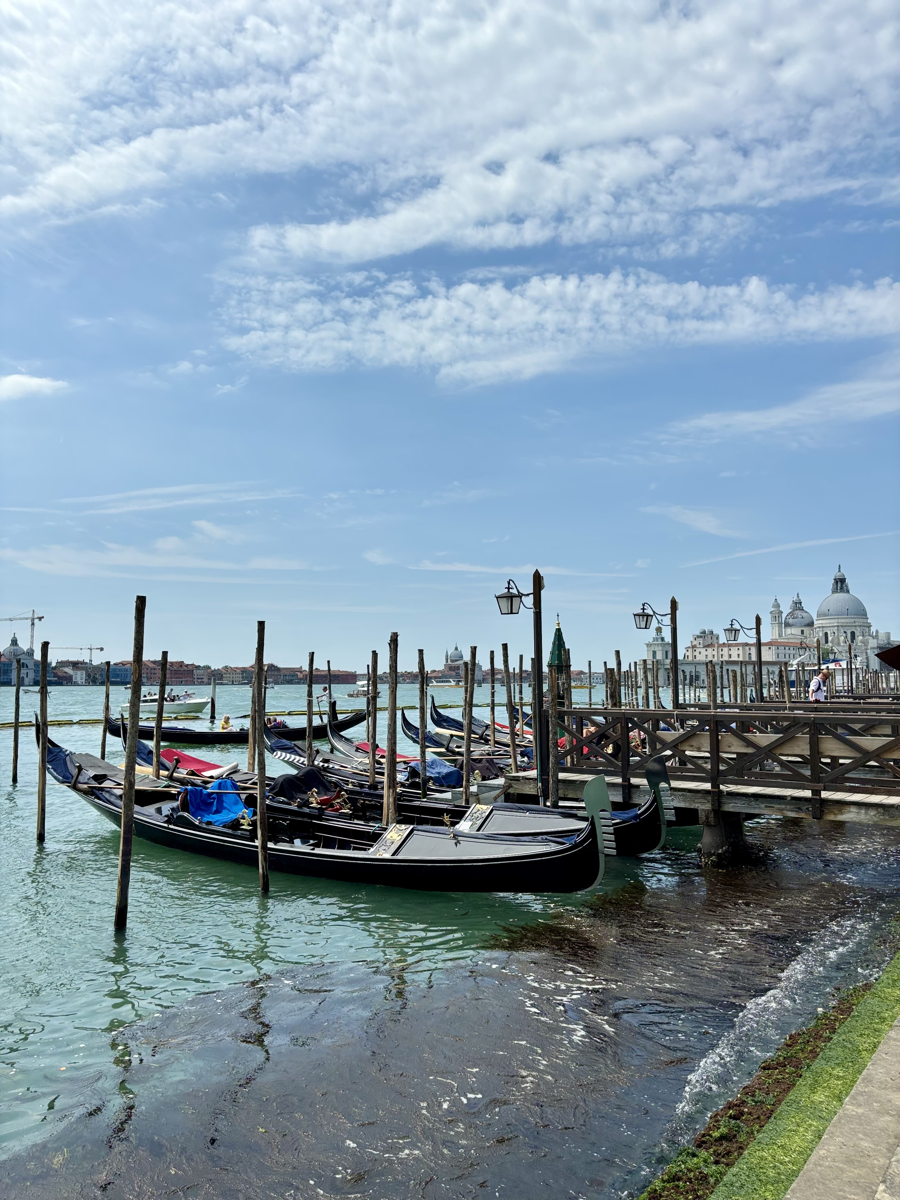 Several gondolas moored at a wooden dock in Venice, Italy, with domed church buildings visible across the water under a partly cloudy sky.