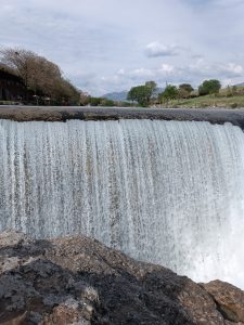 Wide waterfall cascading over a smooth ledge into a river with rocky terrain in the foreground and green trees in the background.
