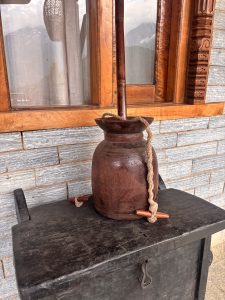 Rustic wooden water pitcher with a carved handle on a dark wooden surface, with stone walls in the background.
