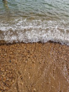Overhead view of a clear ocean wave breaking with white foam onto a wet, golden pebble and shingle beach.