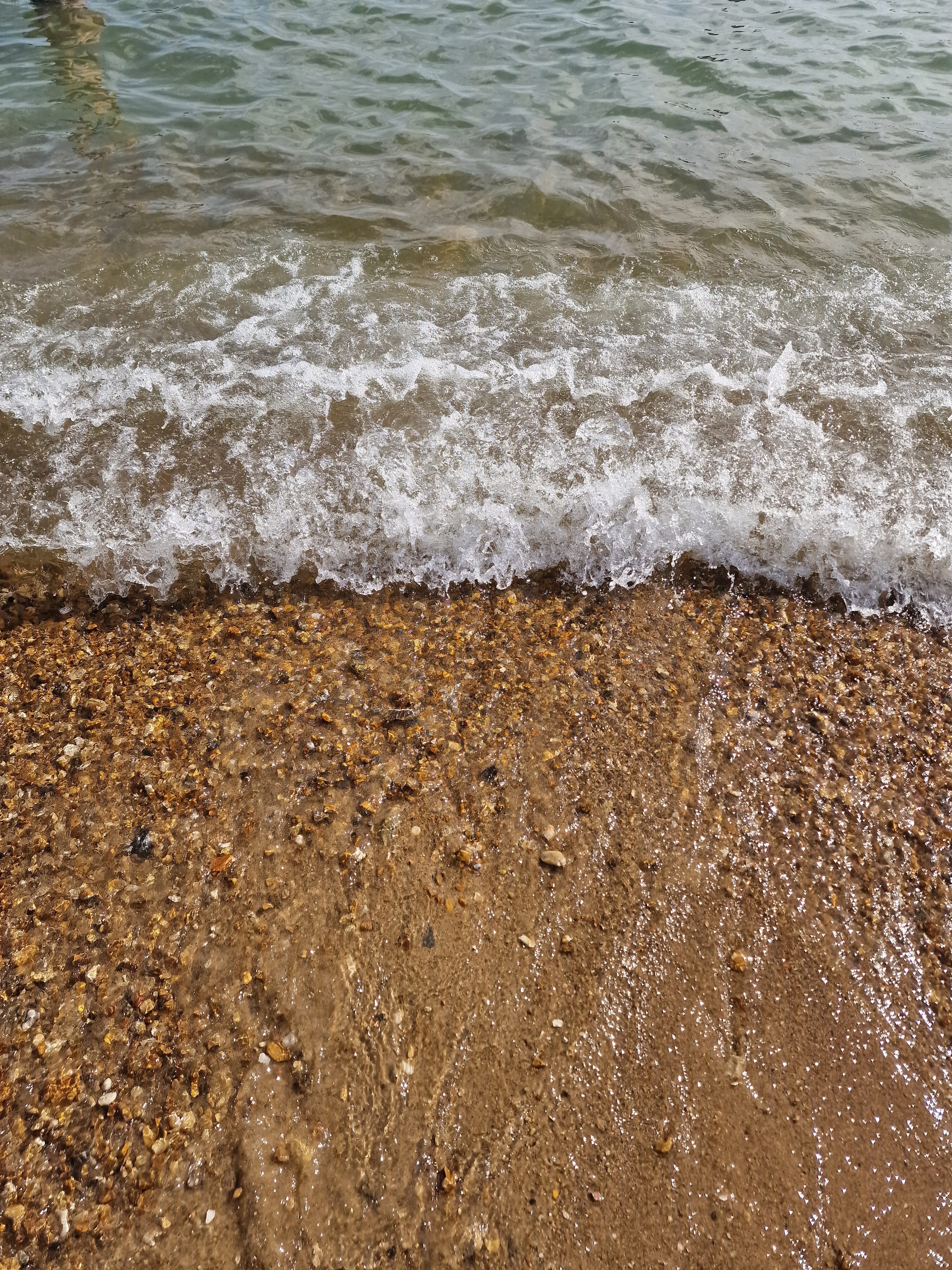 Overhead view of a clear ocean wave breaking with white foam onto a wet, golden pebble and shingle beach.