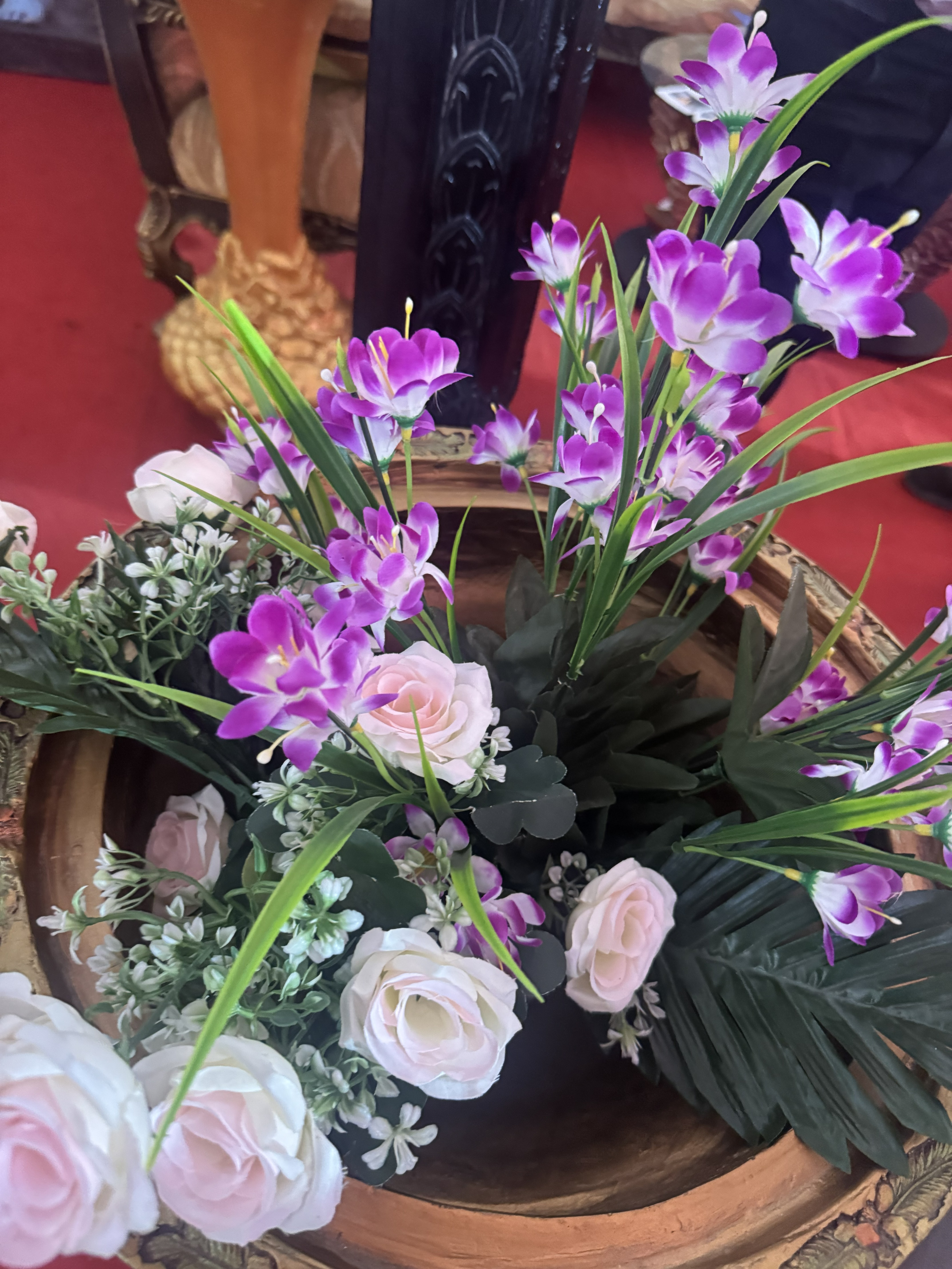 An arrangement of artificial flowers featuring purple blooms, white and pink roses, and green foliage displayed in a large decorative wooden bowl