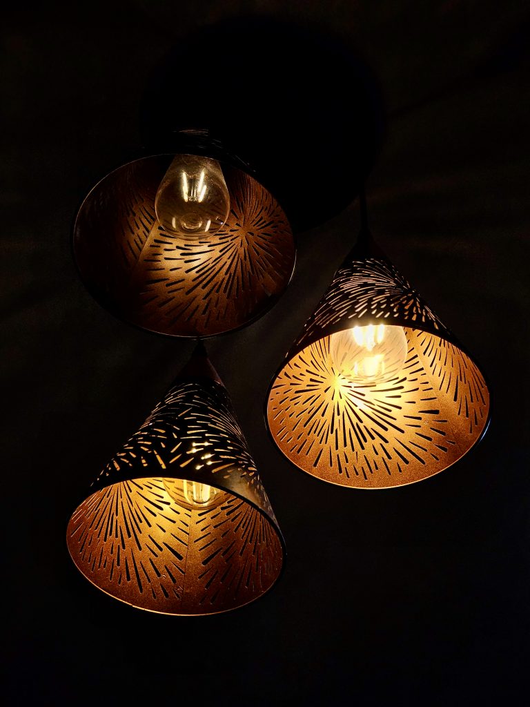 Three decorative pendant lamps glow warmly in the darkness, creating an attractive pattern of light, shadows, and intricate cut-out designs. Location: Ayamkudy, Kottayam, Kerala.