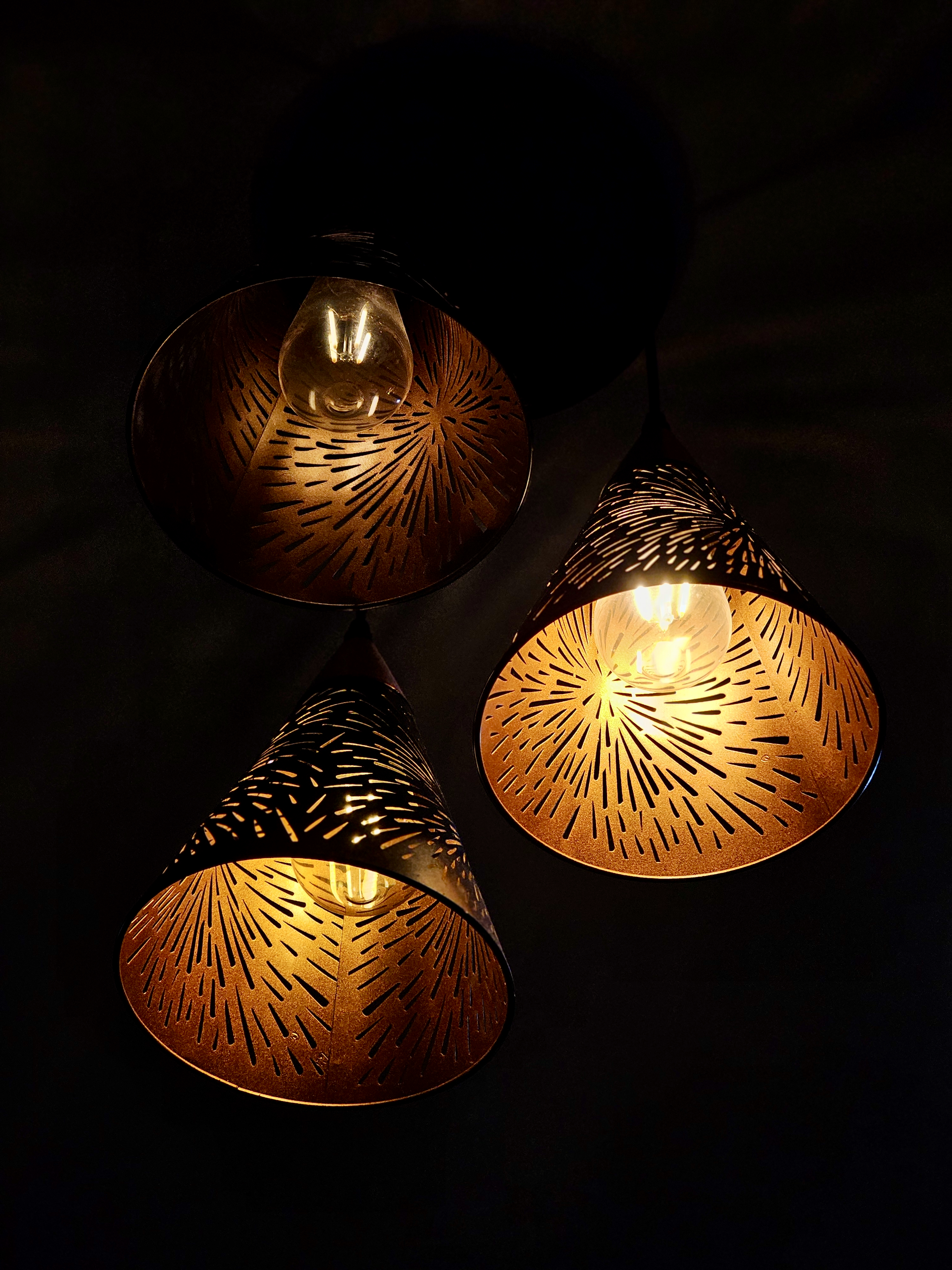 Three decorative pendant lamps glow warmly in the darkness, creating an attractive pattern of light, shadows, and intricate cut-out designs. Location: Ayamkudy, Kottayam, Kerala.
