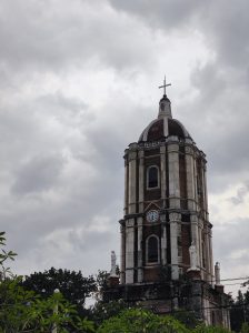 Church clock tower with a domed top, cross, clock face, and statues under a cloudy sky.