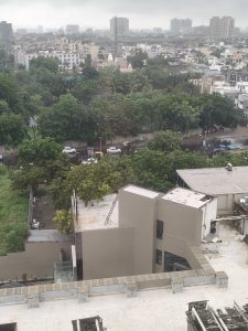 Aerial view of a dense city with rooftops, green trees, roads, parked cars, and buildings under a cloudy sky.