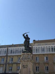 Statue of María Pita holding a raised spear on a stone pedestal in A Coruña, with traditional Galician buildings in the background.