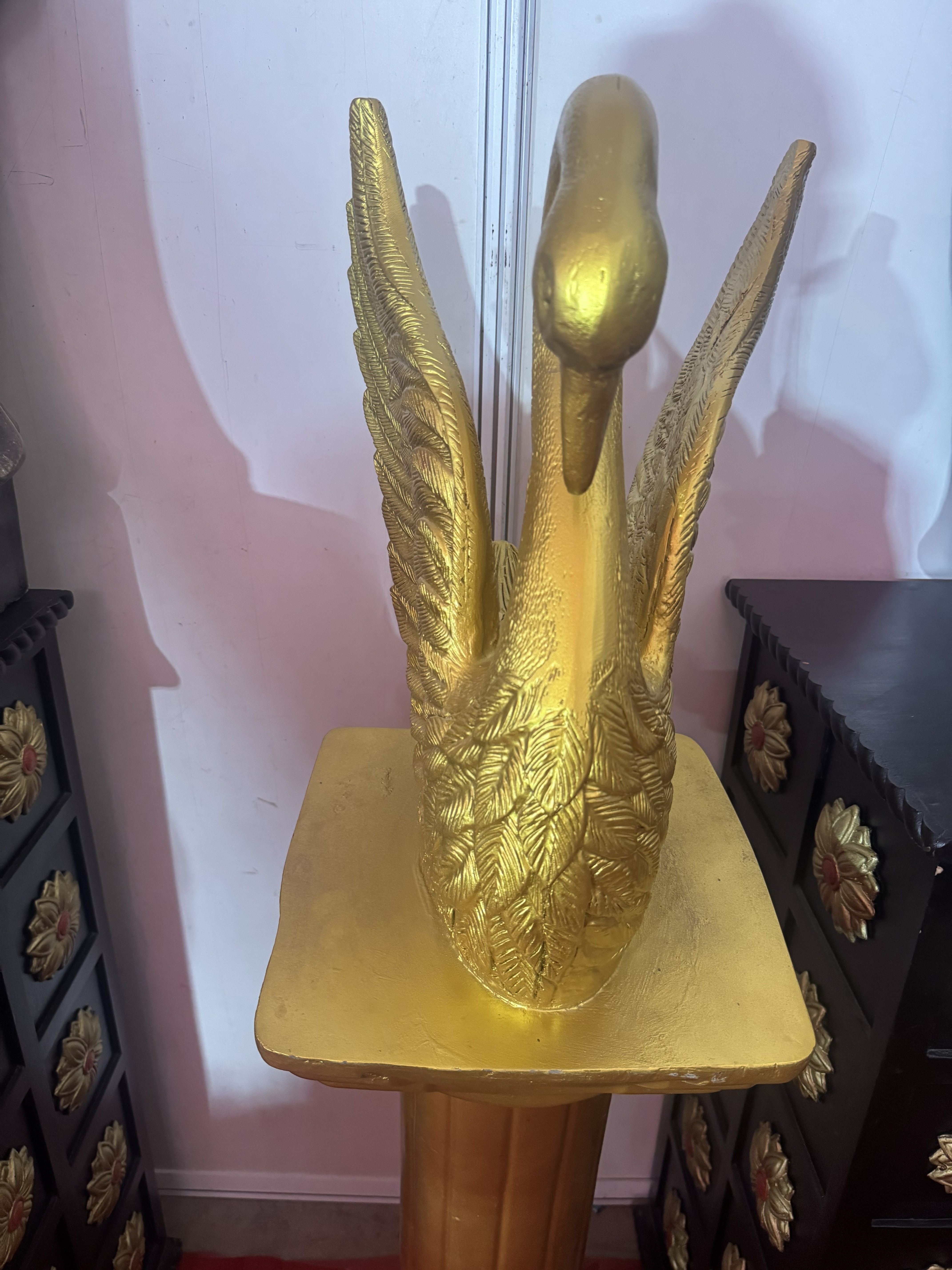 A metallic-gold swan sculpture with outstretched wings mounted on a matching golden pedestal.
