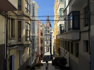 Perspective view of a narrow uphill street leading to A Coruña City Hall, with its reddish dome, colorful residential buildings, and a clear blue sky.