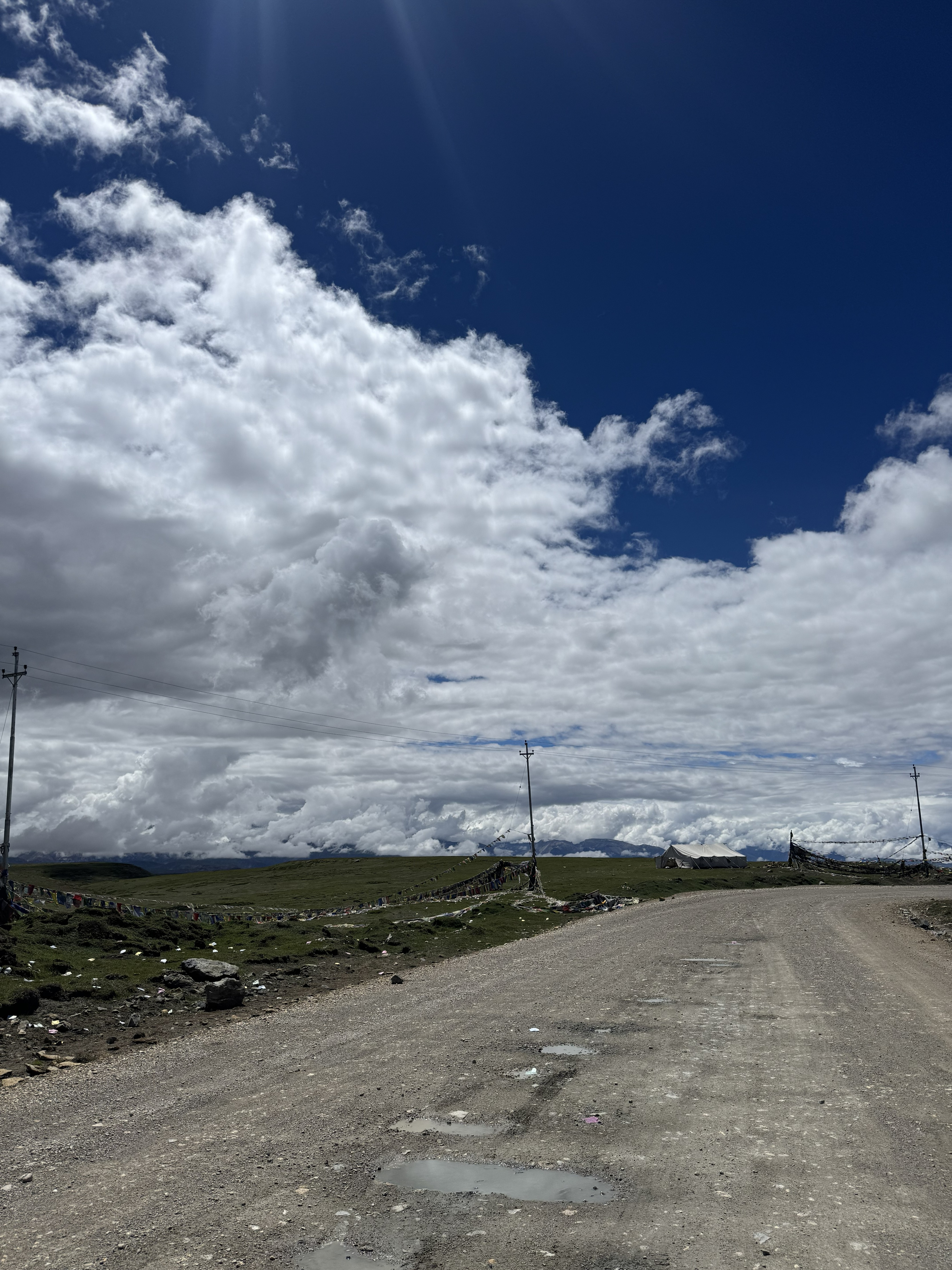 Dirt road with puddles crossing a green high-altitude plain, with prayer flags, utility poles, and a white tent under a cloudy blue sky.