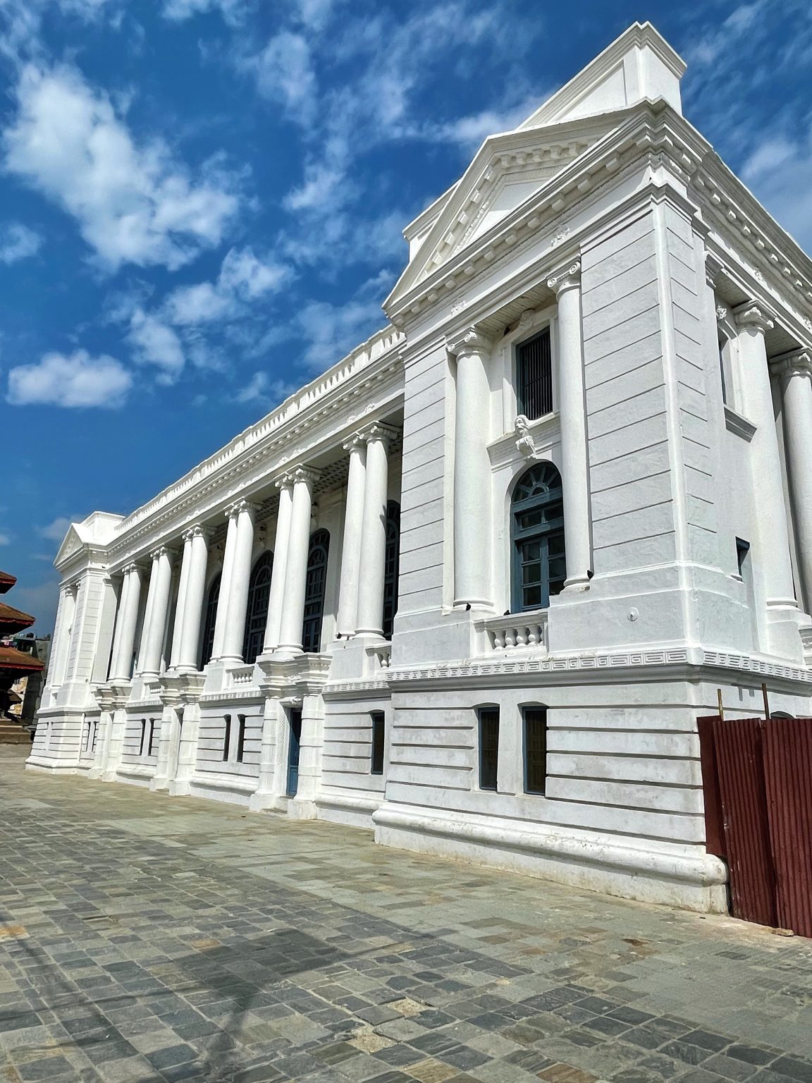 A grand white historic building with tall classical columns and ornate architectural details stands beneath a vivid blue sky with scattered clouds. The stone-paved courtyard adds depth to the architectural scene. Kathmandu, Nepal.