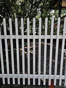 A white picket fence stands in front of a lush garden, with trees, a paved walkway, and part of a house visible behind it. The fence's strong vertical lines create a simple, graphic composition. Location: Ayamkudy, Kottayam, Kerala.