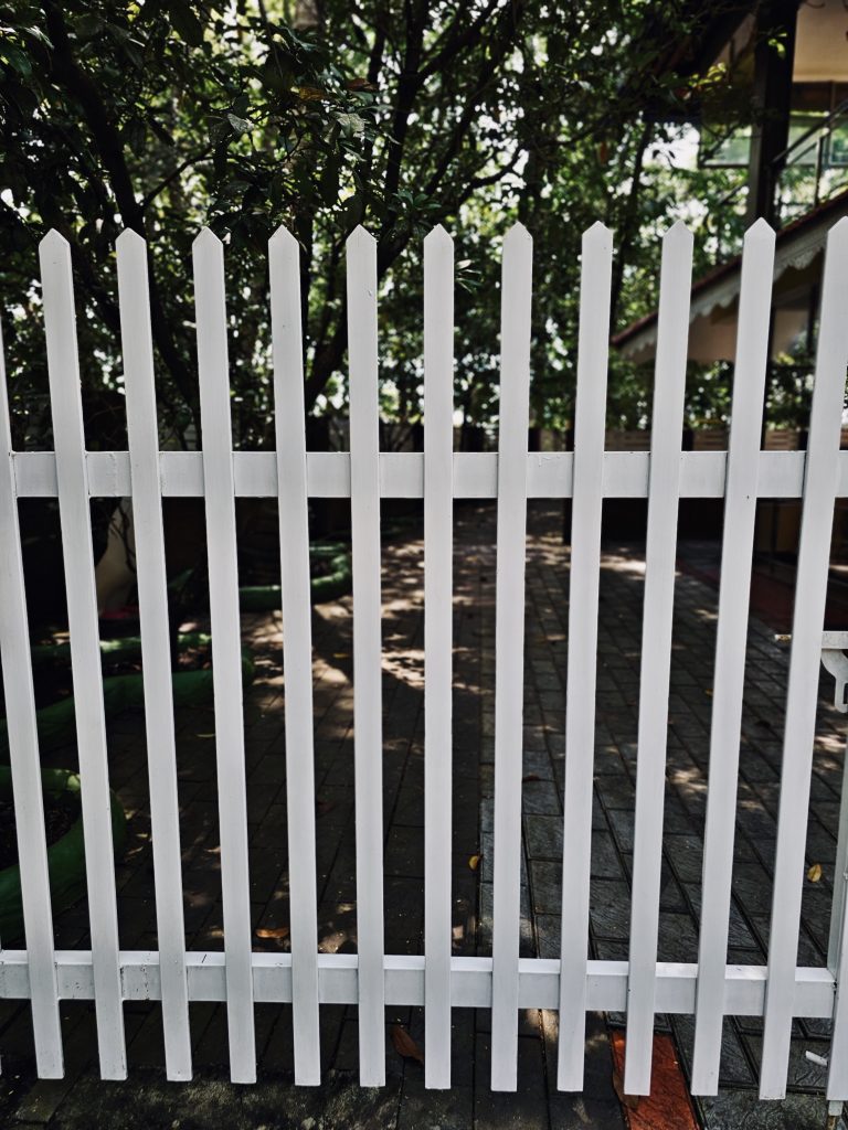 A white picket fence stands in front of a lush garden, with trees, a paved walkway, and part of a house visible behind it. The fence's strong vertical lines create a simple, graphic composition. Location: Ayamkudy, Kottayam, Kerala.