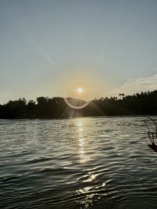 A river at sunset reflects the sun’s rays, creating a shimmering effect on the water’s surface. The silhouetted trees and distant structures add depth to the serene landscape.
