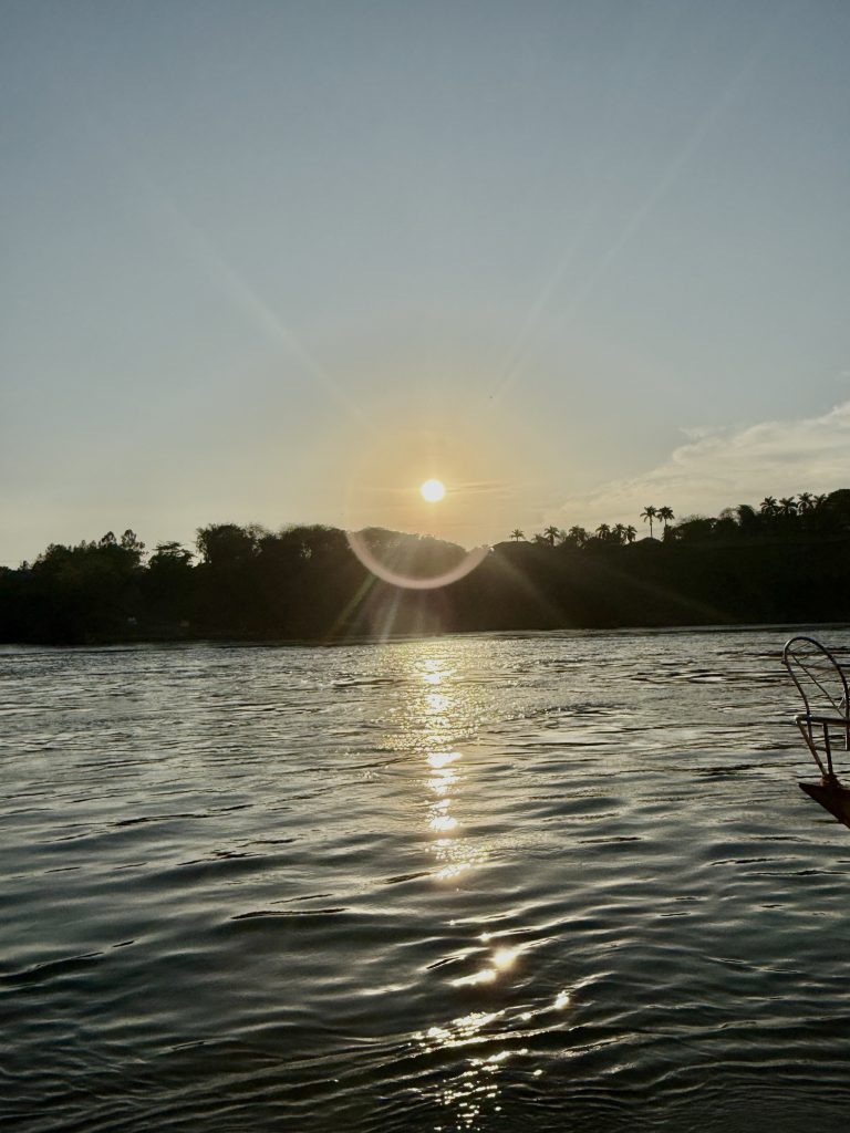 A river at sunset reflects the sun’s rays, creating a shimmering effect on the water’s surface. The silhouetted trees and distant structures add depth to the serene landscape.