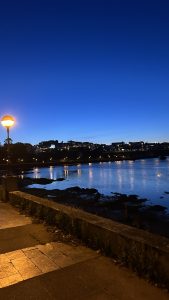 Riverside walkway at sunset with city lights reflected in the water under a deep blue sky.
