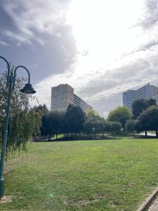 A green park with a lamppost, trees, and a tall modern building under a bright cloudy sky.