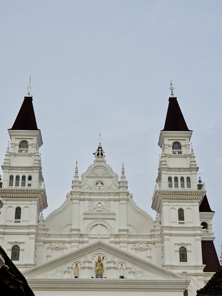 A detailed frontal view of the grand white church, showing its twin towers, decorative façade, stained-glass windows, and ornate architectural details. Location: Ramapuram, Kottayam, Kerala.