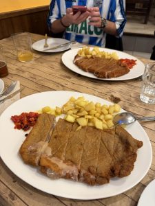 Breaded cachopo cut into strips with melted cheese, served with diced fries and roasted red peppers on a rustic wooden table.