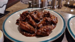 A plate of chicken wings heavily coated in a dark, glossy barbecue sauce, garnished with scattered red pepper flakes and served on a cream-colored plate with a teal rim.
