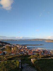 Panoramic view of the coastal village of Fisterra with red-roofed houses, harbor, and beach along the blue ocean bay in Galicia under a clear afternoon sky.