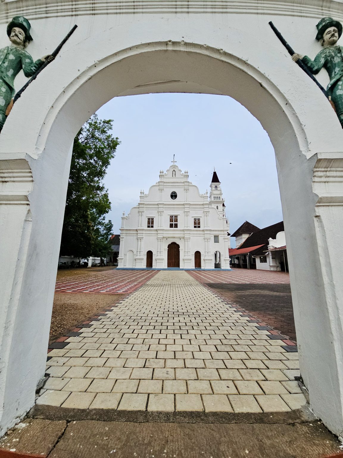A white historic church viewed through a large arched gateway, with two sculpted figures framing the entrance. The paved pathway leads toward the ornate church façade. Location: Ramapuram, Kottayam, Kerala.